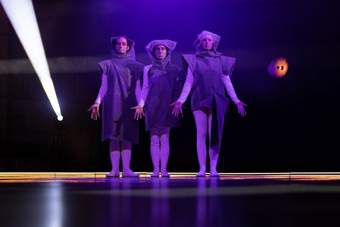 Drei Darsteller stehen nebeneinander auf einer dunklen Bühne in lila Licht. Sie tragen helle, enge Hosen und einen dunklen Umhang mit Kapuze. Ihre Handflächen mit neongrünen Fingerspitzen strecken sie nach vorn.