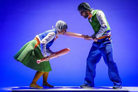 Auf einer blau beleuchteten Bühne stehen sich eine Frau und ein Mann gegenüber. Sie sind blau und grün gekleidet und erinnern mit ihren Perücken an Legofiguren. Sie kämpfen mit aufblasbaren Baseballschlägern. 