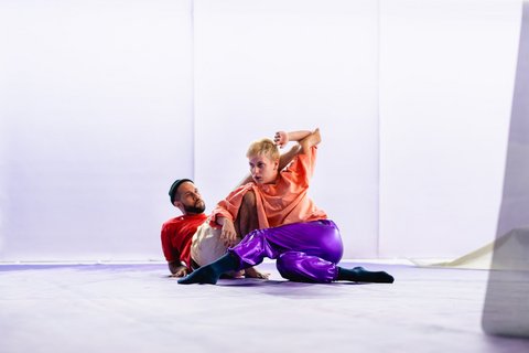 Eine Tänzerin und ein Tänzer in farbigen Outfits liegen ineinander verschränkt auf dem Boden einer weißen Bühne.