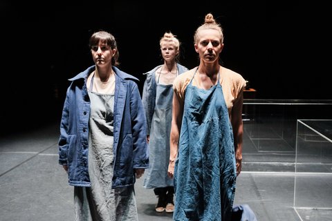 Drei Frauen in blauen und grauen Schürzen stehen nebeneinander auf einer Bühne und blicken starr in die Kamera.