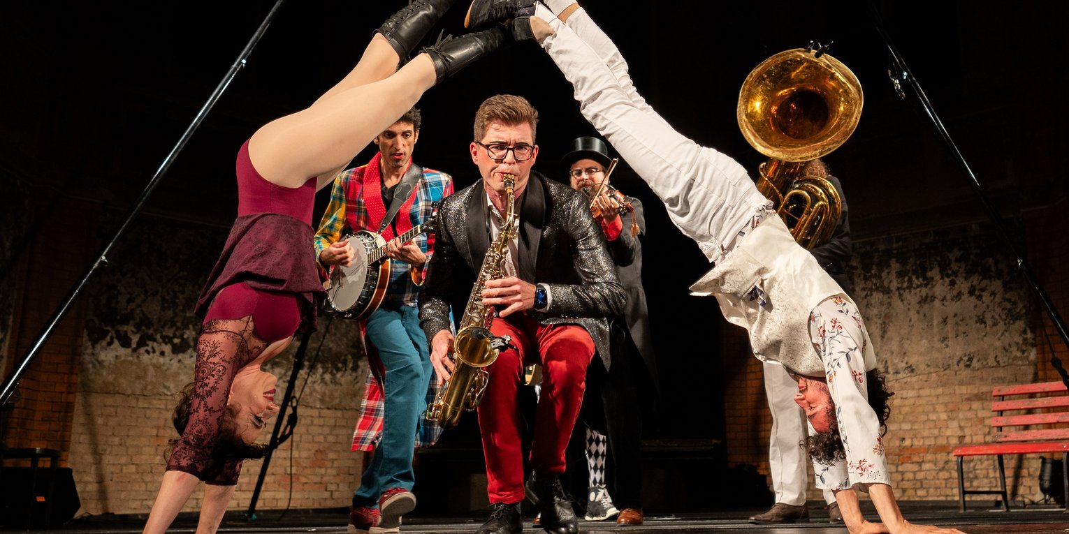 Eine Akrobatin und ein Akrobat stehen sich im Handstand gegenüber, ihre Füße berühren sich. Hinter ist ein Saxophonist und weitere Musiker auf der Bühne zu sehen.