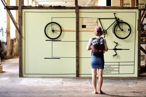 Eine Frau betrachtet eine großflächige, in Holzbalken gerahmte Arbeit, auf der die Bauteile eines schwarzen Fahrrads im Seitenprofil und in der Vogelperspektive montiert sind. 