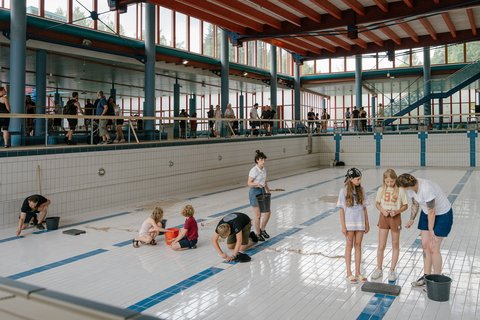 In einem blau-weiß gefliesten Hallenbad putzen einige Erwachsene und Kinder im leeren Becken die Fliesen am Boden.