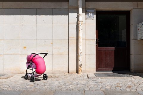 Ein rosa Kinderwagen steht auf einer Kopfsteinpflasterstraße vor einem Hauseingang. Aus dem Kinderwagen quillt pinken Schaum. 
