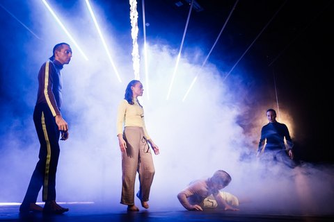 Auf einer Bühne stehen vier Performer in statischen Posen, hinter ihnen steigt mannshoch Nebel aus einer Nebelmaschine auf. Ein heller, bläulicher Lichtkegel beleuchtet die Bühne.