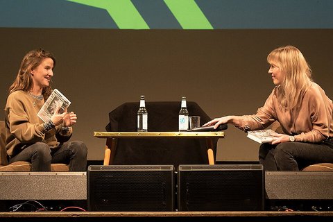Auf einer Bühne sitzen sich zwei Frauen auf Sesseln an einem Tisch gegenüber und unterhalten sich.