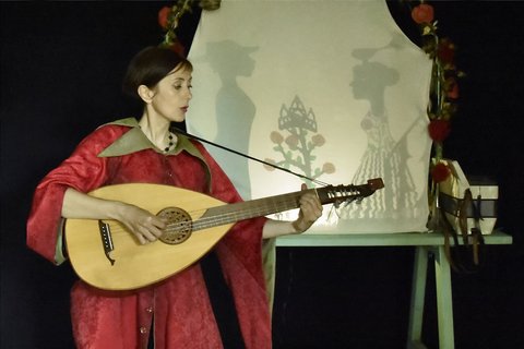 Eine Frau in einem roten Kleid spielt eine Laute, im Hintergrund ist ein Schattenspiel zu sehen.