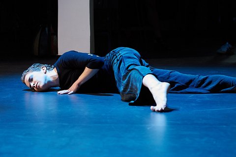 Auf einem beleuchteten Tanzboden liegt bäuchlings eine Performerin in einem schwarzen Outfit.