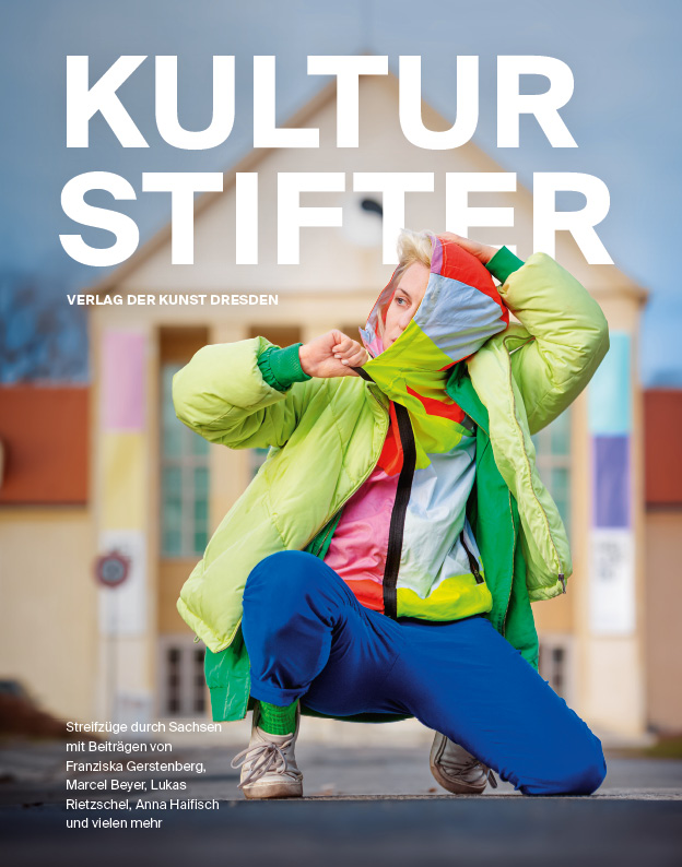 Das Cover des Bookzine ist zu sehen. Es zeigt eine sich hinhockende Frau in blauer Hose und bunter Jacke. Im Hintergrund ist das Festspielhaus Hellerau zu sehen.