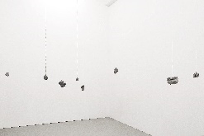 In einem weißen Ausstellungsraum hängen rundliche, graue Betonobjekte an Metallketten von der Decke. 