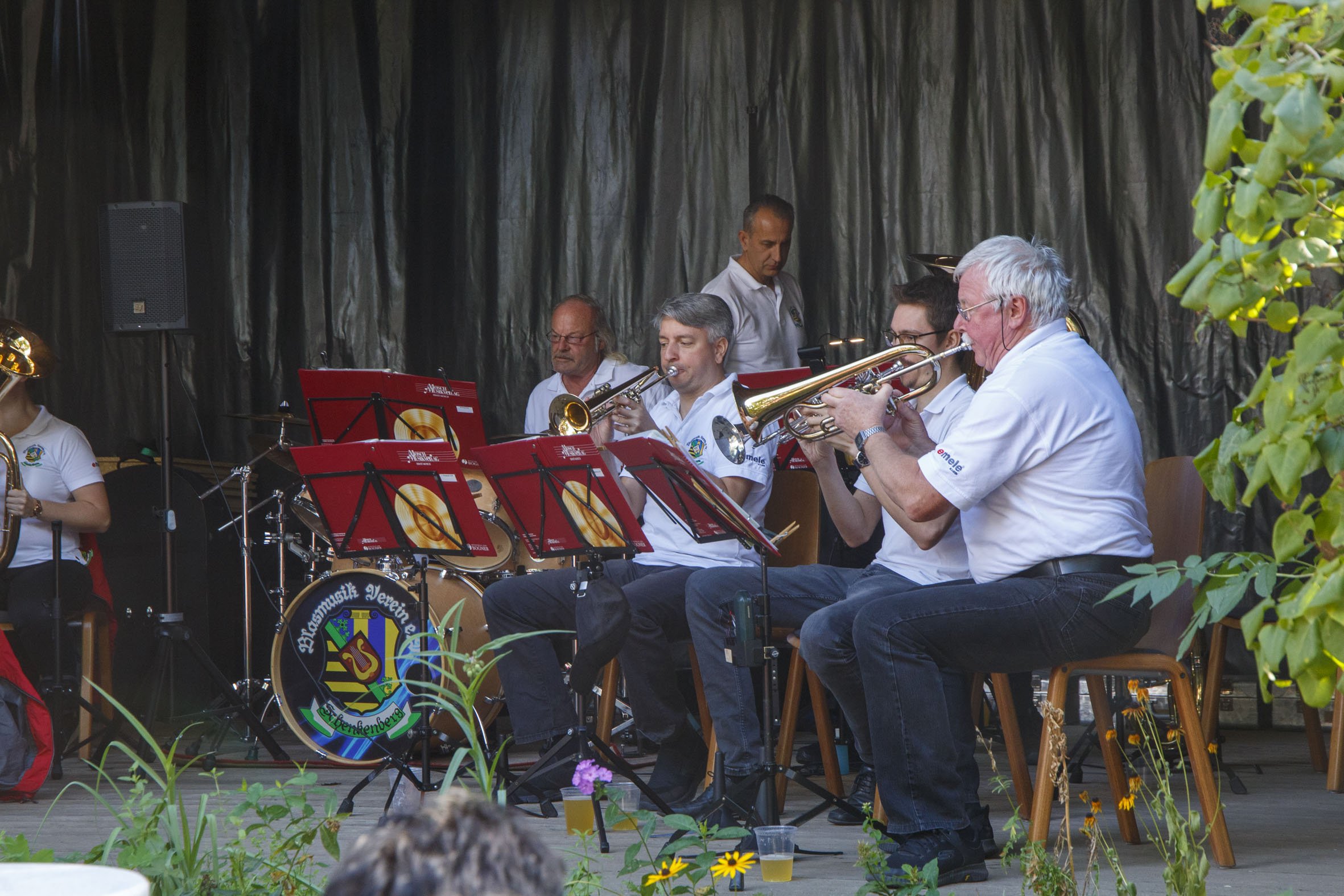 Ein Blasorchester spielt im Freien auf einer Holzbühne vor einer Blumenwiese.