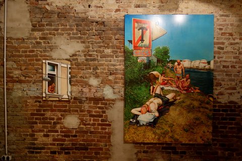 An einer Ziegelsteinwand hängen zwei Gemälde. Auf dem einen ist ein Mann zu sehen, der sich aus dem Fenster lehnt und auf dem anderen sind Badende an einem See abgebildet. 