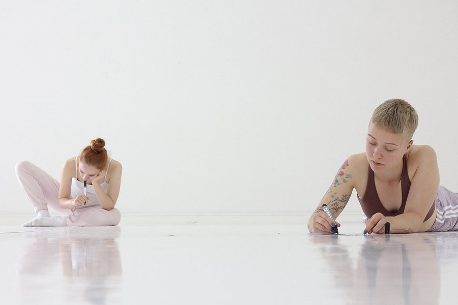 In einem weißen Raum liegen bzw. sitzen zwei Frauen in einiger Entfernung nebeneinander auf dem Boden. Jede schreibt mit Filzstift etwas auf ein Blatt Papier.