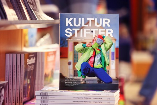 Das Magazin Kulturstifter aufgestellt in einer Buchhandlung auf einem Bücherstapel, auf dem Cover ist eine farbenfroh angezogene Tänzerin vor dem Festspielhaus Hellerau zu sehen.