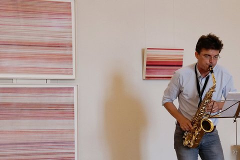 Ein Mann spielt vor einer Wand mit gestreiften Bildern Saxofon. 
