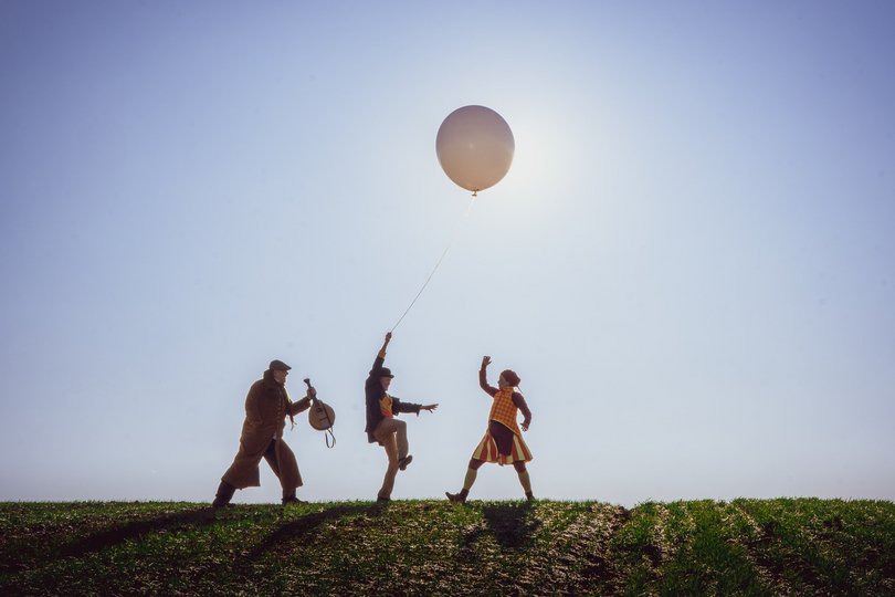Auf einem Feld sind drei kostümierte Personen vor einem wolkenlosen Himmel zu sehen. Während einer von ihnen eine Mandoline hält, lässt ein anderer einen großen Ballon über sich schweben. 