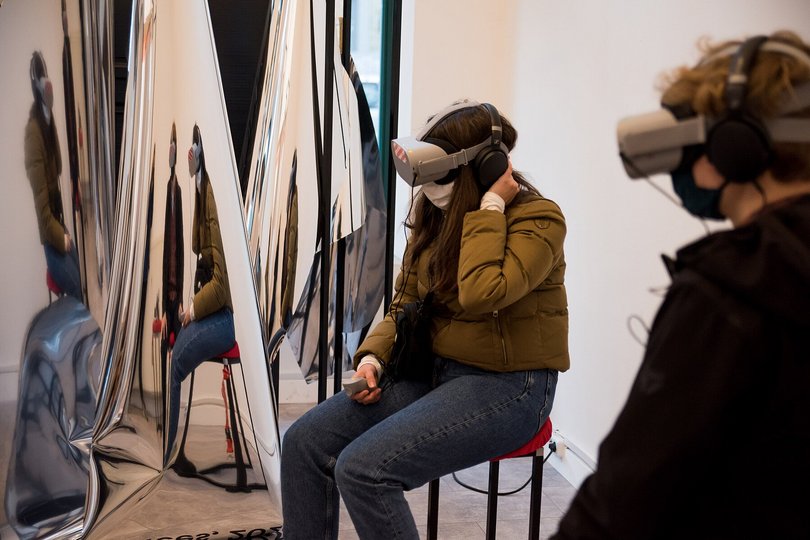 Zwei Menschen tragen VR-Brillen und Kopfhörer und sitzen nebeneinander vor einer Spiegel-Skulptur. 