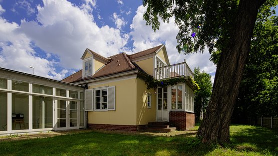 Ein gelbes Haus mit einer Dachgaube, weißen Fensterläden und einem gläsernen Wintergarten wird von Ranken und einer Ziegelsteinverzierung gerahmt.