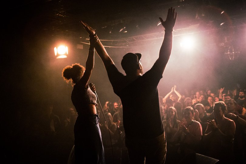 Die Rückenansicht von einem Mann und einer Frau, die sich bei den erhobenen Händen fassen.  Sie stehen auf einer Bühne vor applaudierendem Publikum und werden von Bühnenleuchten angestrahlt. 