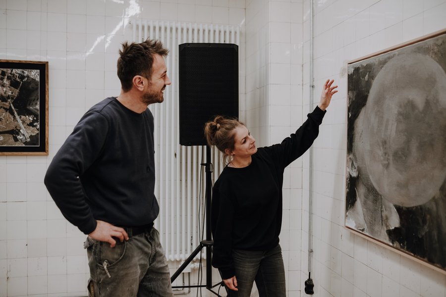 Zwei Personen stehen vor einem großformatigen Kunstwerk an einer weißen Wand, eine Person zeigt mit erhobenem Arm darauf.