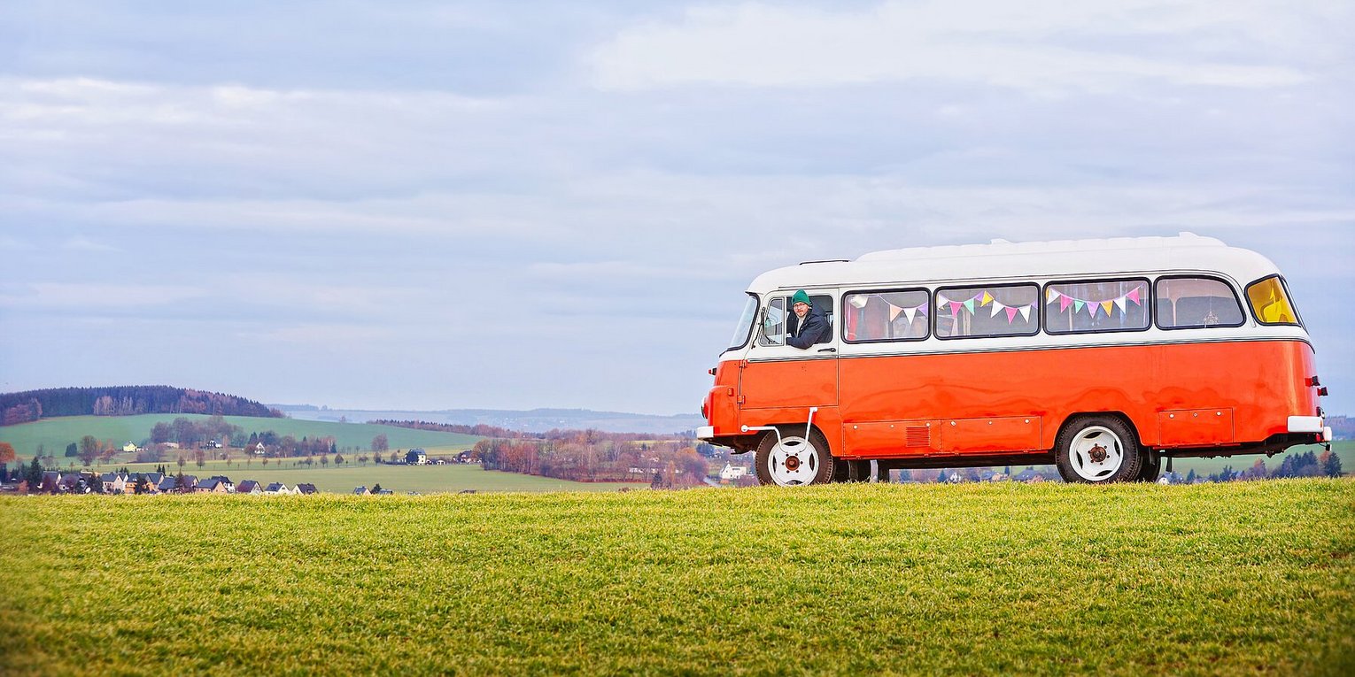 Auf einem grünen Hügel steht ein orangenfarbener DDR-Oldtimer-Bus.