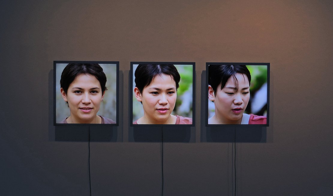 Drei an einer Wand hängende Bildschirme zeigen dieselbe Frau mit drei verschiedenen Gesichtsausdrücken.