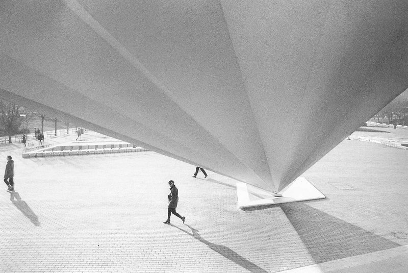 Schwarz-weiß Fotografie von Menschen, die über einen Platz laufen. Durch die obere Hälfte des Fotos läuft über die gesamte Breite eine Stahlstrebe, die zu einem Punkt verläuft.