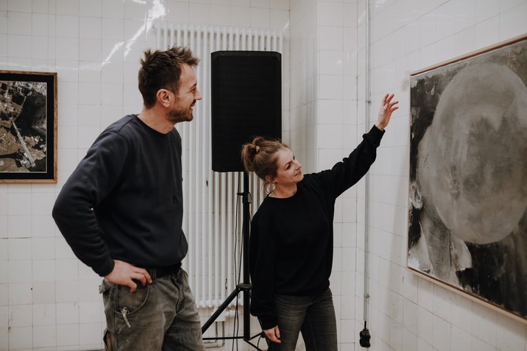Pressebild im Format JPG öffnen: Zwei Personen stehen vor einem großformatigen Kunstwerk an einer weißen Wand, eine Person zeigt mit erhobenem Arm darauf.