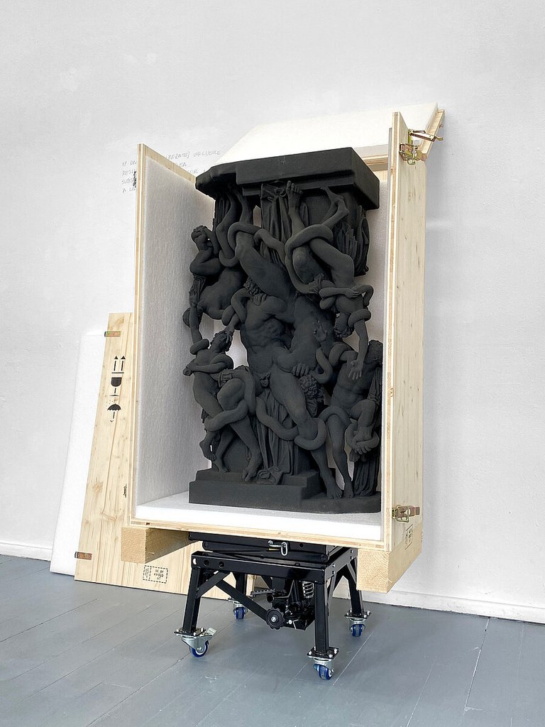 In einer hölzernen Transportkiste steht eine schwarze Skulptur, die aus einer Säule und mehreren ineinander verschlungenen Körpern besteht.