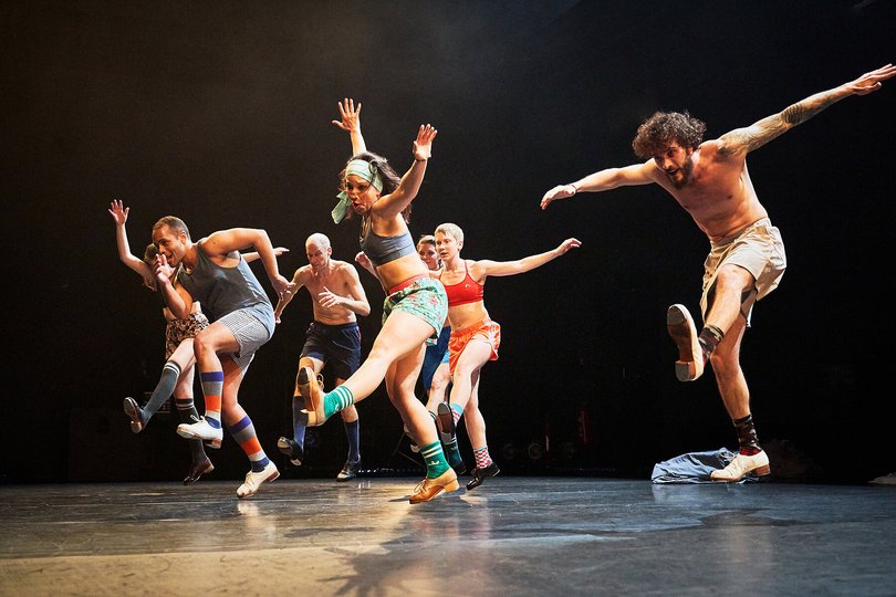 FOLK FICTION, Sebastian Weber Dance Company, Foto: Tom Dachs Auf einer schwarzen Bühne springt eine Gruppe Tänzer mit ausgestreckten Beinen und Armen gleichzeitig nach vorn.