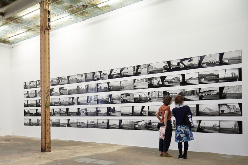 In einem Ausstellungsraum stehen zwei Frauen vor einer schwarz-weißen Fotoreihe. Eine weitere Person steht vor drei farbigen Gemälden.