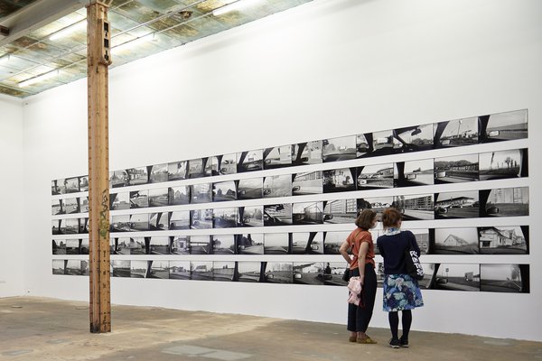 In einem großen Raum mit Stahlträgern stehen zwei Frauen vor einer Wand mit schwarz-weiß Fotos.  