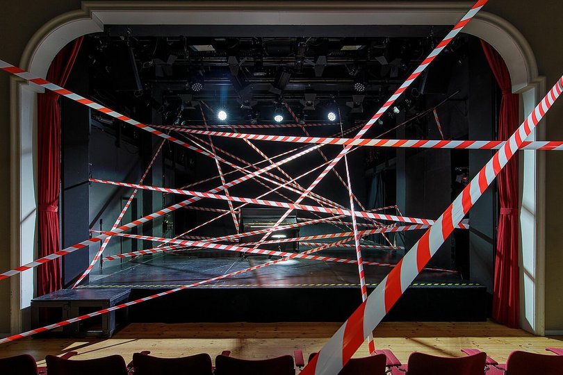 Eine große Bühne mit davorstehenden Stühlen wird quer durch den Raum von rot-weißem Absperrband durchzogen.