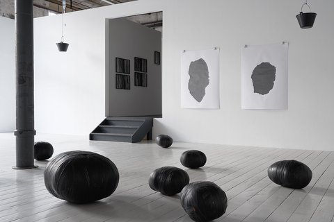 In einem Ausstellungsraum liegen verschieden große, mit schwarzem Klebeband gebundene Bündel auf grauem Dielenboden. An der Wand hängen Drucke und Zeichnungen in schwarz-weiß.