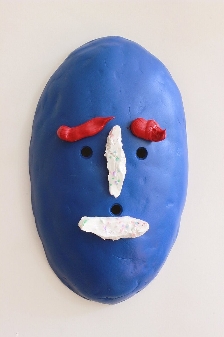 Eine blaue Maske mit drei Löchern für Augen und Ohren. Als Augenbrauen ist großzügig und unregelmäßig eine rote Farbschicht aufgetragen. 