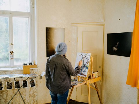 In der Ecke eines Zimmer steht eine Person und malt an einem Gemälde auf einer Staffelei.