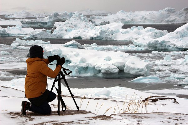 Eine Frau hockt in einer Eislandschaft hinter einer Kamera mit Stativ.