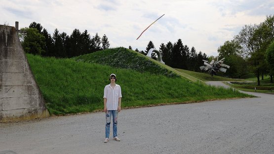Peter Piek steht mit weißen Hemd, Jeans und weißer Cappy auf einer Kiesstraße. Hinter ihm ist ein kleiner Hügel und seine Skulptur.