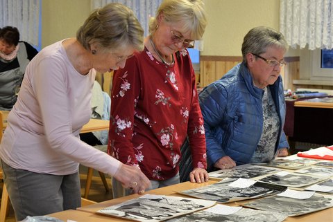 Drei älterer Frauen schauen sich schwarz-weiß Fotografien an, welche auf einem Tisch liegen.
