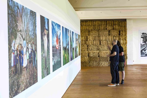 Zwei Männer betrachten ein Bild in einer Kunstausstellung. Im Hintergrund ist eine Wand aus Heuballen zu sehen.