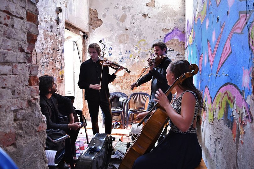 In einem engen Raum mit unverputzten Wänden stehen wir festlich gekleidete Musiker im Kreis und halten ihre Streichinstrumente in den Händen.