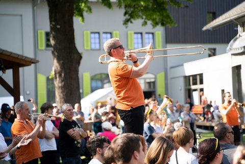 In schwarz und orange gekleidete Blechbläser spielen ein Konzert in einem Innenhof und inmitten von Publikum. Ein Spieler steht erhöht auf einem Podest. 