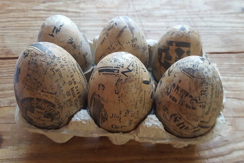 Auf einem Holztisch steht ein Eierkarton mit sechs Eiern, welche mit Zeitungspapier beklebt sind. 