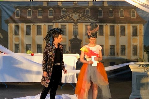 Zwei Schauspieler in Kostümen auf einer Bühne, im Hintergrund eine bedruckte Leinwand. 