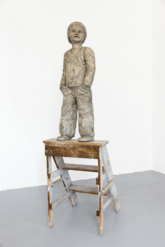 Auf einem Holztritt steht die Skulptur eines Kindes aus Pappmaché.