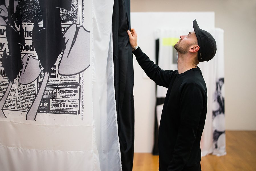 Ein Mann berührt einen schwarz bedruckten Vorhang, welcher von der Decke hängt, und blickt an ihm hinauf. 