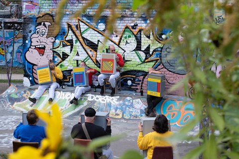 Am Rand einer flachen, mit Graffiti besprühten Halfpipe sitzen Personen auf Stühlen im Kreis und halten buntbemalte Kästen auf ihrem Schoß.
