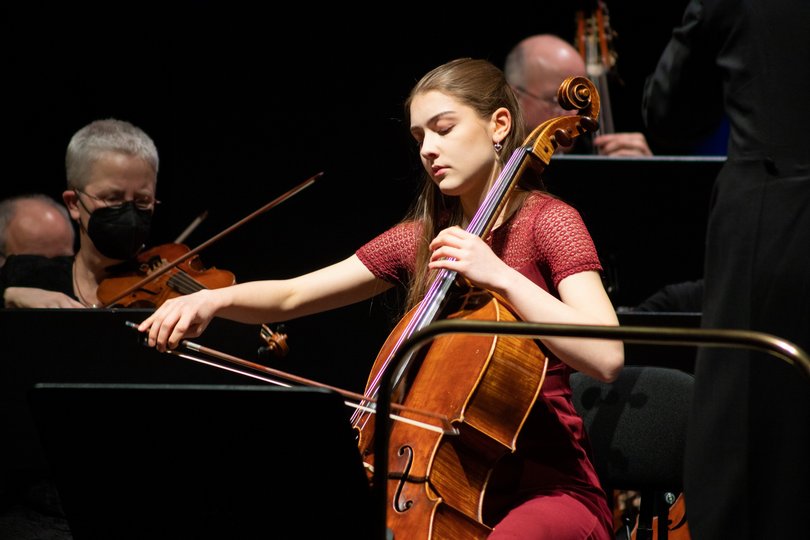 Eine junge Frau in einem roten Kleid spielt während eines Orchesterkonzerts Cello. Sie konzentriert sich auf die Musik, ihre Augen sind geschlossen. 