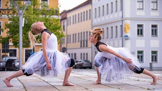 Zwei Tänzerinnen in einer Fußgängerzone. Sie tragen weiße Tüllkleider und stehen sich im Ausfallschritt gegenüber.