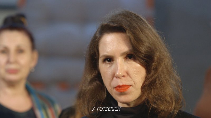 Standbild eines Videos, auf dem eine Frau mit braunen Haaren und rotem Lippenstift zu sehen ist. Das Wort "Fotzerich" ist als Untertitel eingeblendet.